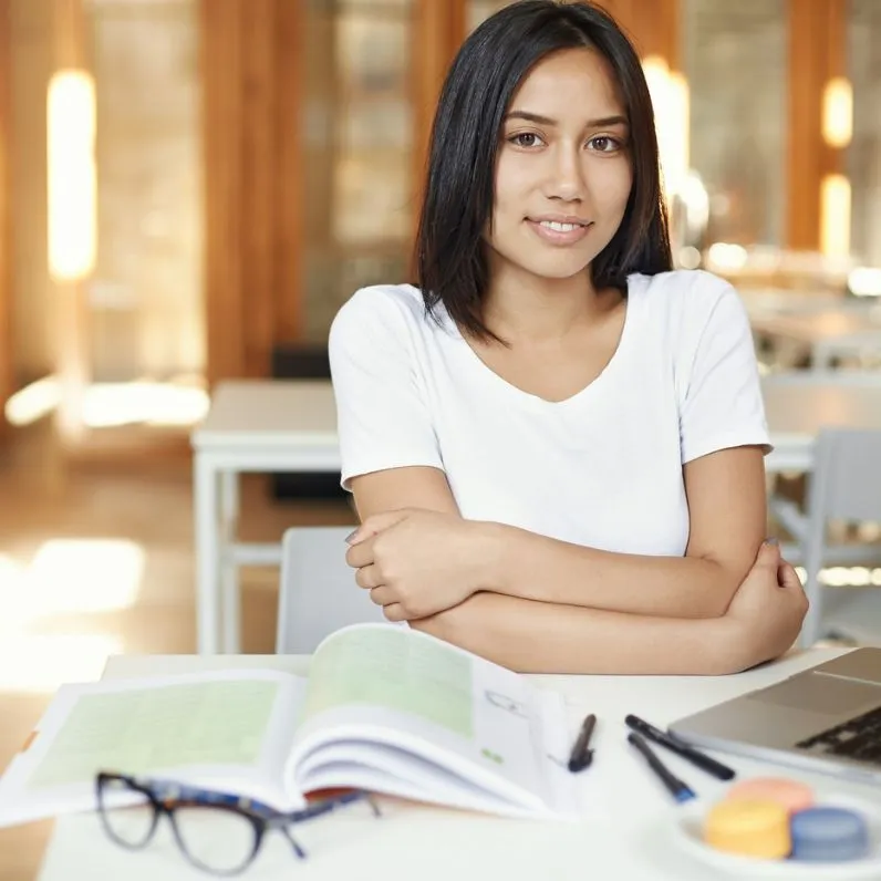 Young woman sitting at a desk with an open book, pens, and glasses in front of her, smiling and looking at the camera.