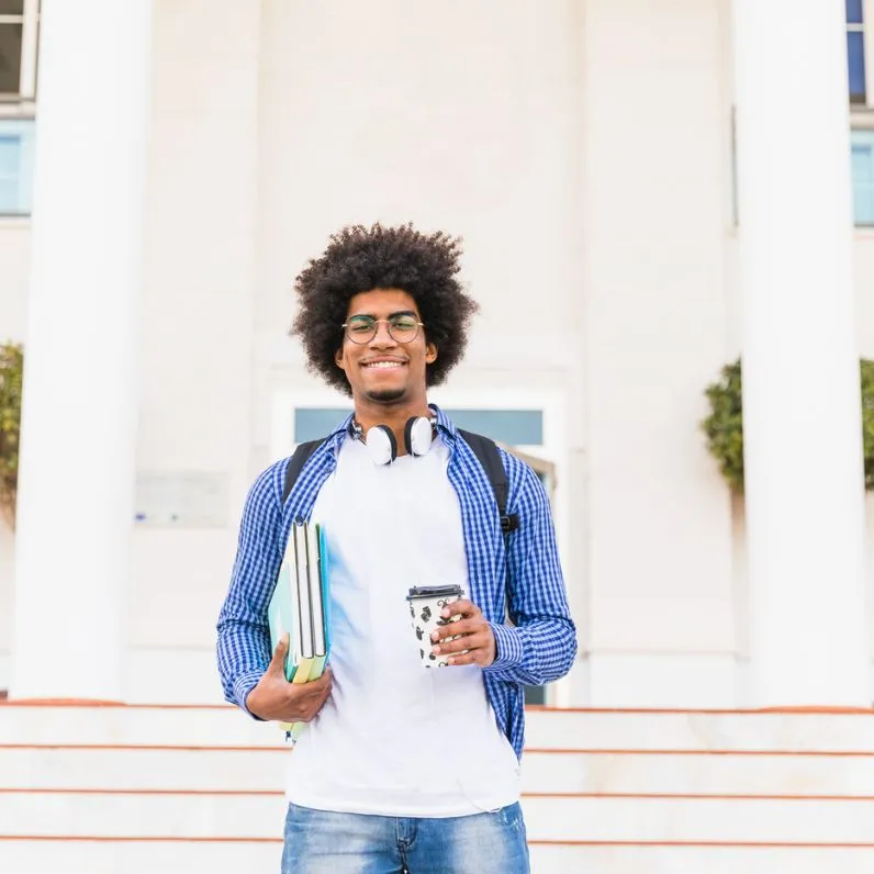 A smiling student in a checkered blue shirt, holding books and a coffee cup, stands outside a building with white columns.